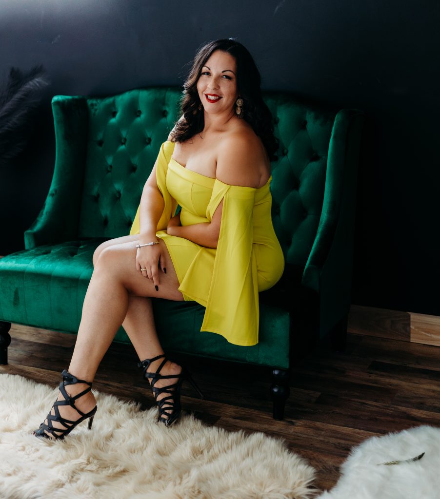 Dr. Alex sitting in a green chair with a yellow dress, black high heels, red lipstick, dark hair, furry white carpet, smiling.